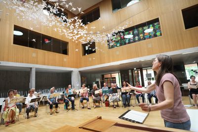 Chorprobe in einem hellen Raum mit Oberlicht. Im Vordergrund ist die Chorleiterin zu sehen, die gerade dirigiert. Im Hintergrund sitzen die Sängerinnen und Sänger halbkreisförmig in zwei Reihen und singen.