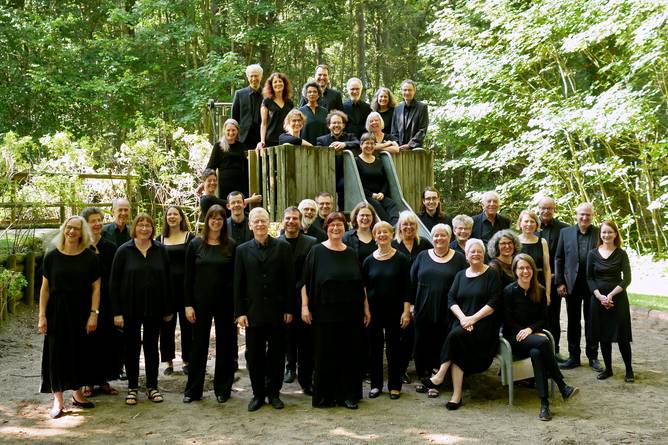 Der Südwestdeutsche Kammerchor in schwarzer Konzertkleidung auf einem Spielplatz im Wald. Einige Personen haben sich auf einem kleinen Holzturm aufgestellt, andere stehen oder sitzen davor. Alle blicken fröhlich in die Kamera.