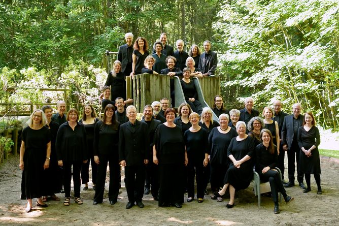 Gruppenfoto vom Chor in schwarzer Konzertkleidung auf einem Spielplatz im Wald. Einige Sänger und Sängerinnen haben sich auf einem kleinen Holzturm mit Rutsche aufgestellt, andere stehen oder sitzen davor. In der vordersten Reihe sitzt die Dirigentin. Alle blicken fröhlich in die Kamera.