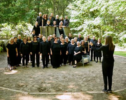 Gruppenfoto vom Chor in schwarzer Konzertkleidung auf einem Spielplatz im Wald. Einige Sänger und Sängerinnen haben sich auf einem kleinen Holzturm mit Rutsche aufgestellt, andere stehen davor. Sie singen gerade und sehen dabei auf die Dirigentin, die mit dem Rücken zur Kamera im Vordergrund steht.