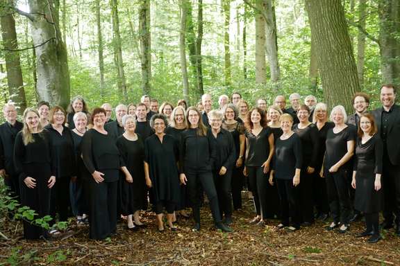 Der Südwestdeutsche Kammerchor steht in schwarzer Konzertkleidung im Wald. Alle blicken fröhlich in die Kamera.