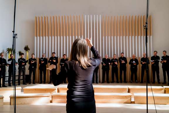 Judith Mohr dirigiert den Südwestdeutschen Kammerchor während eines Konzerts. Sie steht mit dem Rücken zur Kamera im Vordergrund. Im Hintergrund sind etwa sechzehn männliche Chorsänger in schwarzer Konzertkleidung zu sehen, die in einer Reihe stehen.