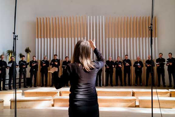Judith Mohr dirigiert den Südwestdeutschen Kammerchor während eines Konzerts. Sie steht mit dem Rücken zur Kamera im Vordergrund. Im Hintergrund sind etwa sechzehn männliche Chorsänger in schwarzer Konzertkleidung zu sehen, die in einer Reihe stehen.
