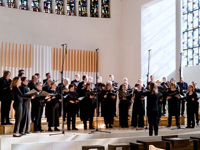 Der Südwestdeutsche Kammerchor während eines Konzerts in St. Petrus, Tübingen. Der moderne Kirchenraum wird durch ein großes, buntes Glasfenster auf der rechten Seite erleuchtet. Das Licht fällt von schräg oben auf die Sängerinnen und Sänger, die schwarze Konzertkleidung tragen.