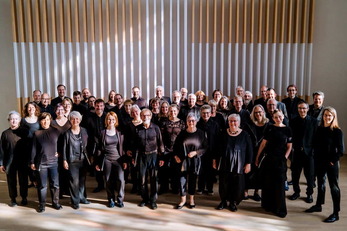 Gruppenbild des Südwestdeutschen Kammerchors Tübingen. Die Sängerinnen und Sänger stehen in einem hellen Kirchenraum in mehreren Reihen vor einer Holzlamellenwand, die ein Kreuz symbolisiert. Sie tragen schwarze Konzertkleidung und blicken fröhlich in die Kamera.