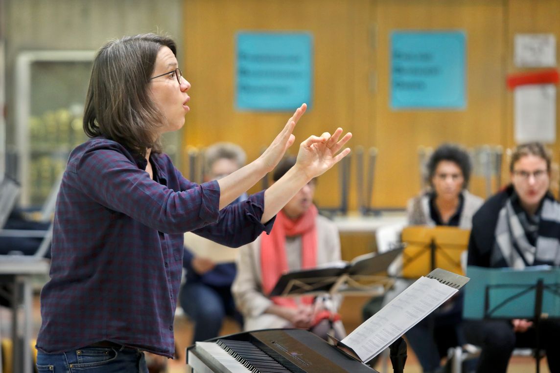 Die Dirigentin seitlich im Profil in leitender Gestik und Mimik vor einem E-Piano und aufgeschlagenen Noten. Im Hintergrund sitzen vier Sängerinnen mit Notenmappen, die der Dirigentin aufmerksam folgen. Das Bild zeigt die dynamische Interaktion zwischen Chorleitung und Sänger:innen während einer Probe.