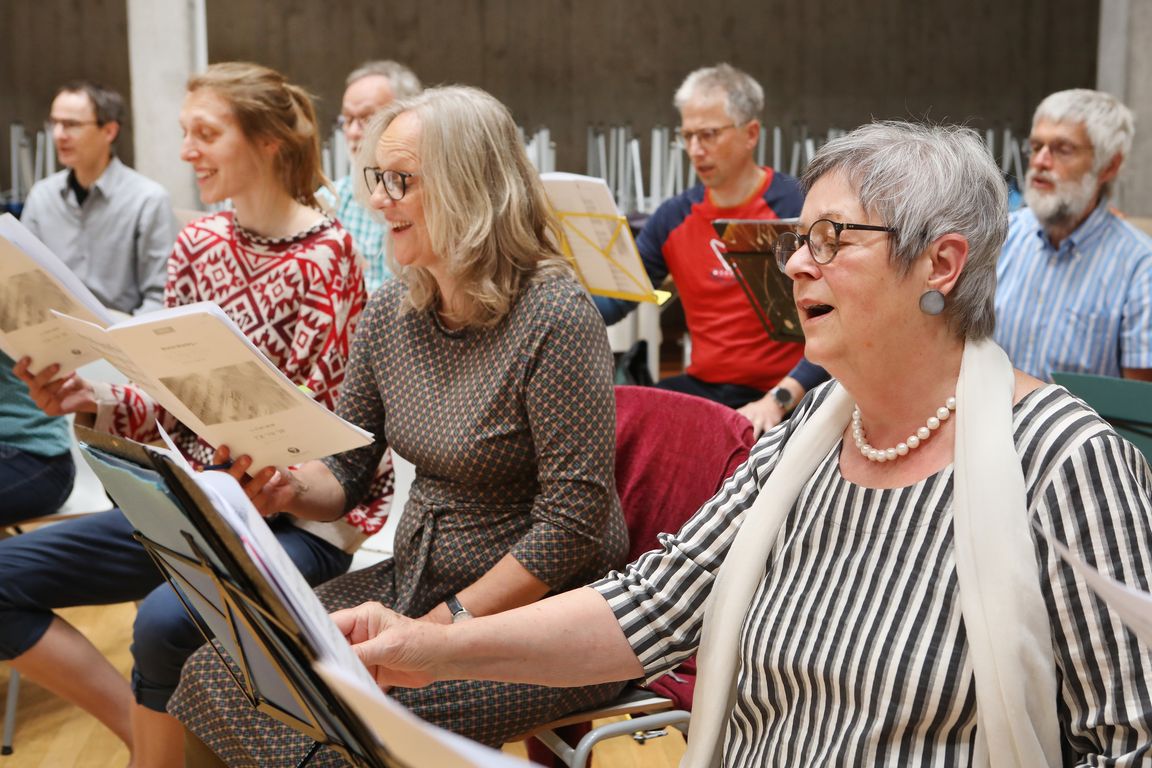Mehrere Chorsängerinnen und -sänger unterschiedlichen Alters sitzen neben- und hintereinander im Probenraum und singen aus aufgeschlagenen Noten. Die Gruppe ist konzentriert, die Körperhaltung aufrecht, der Blick auf die Partituren gerichtet.