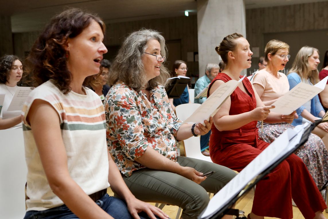 Eine Gruppe von Sängerinnen sitzt im Halbkreis und hält Notenblätter in den Händen. Die Personen sind verschiedenen Alters und wirken engagiert, während sie gemeinsam singen. Im Hintergrund sind weitere Chormitglieder zu erkennen.