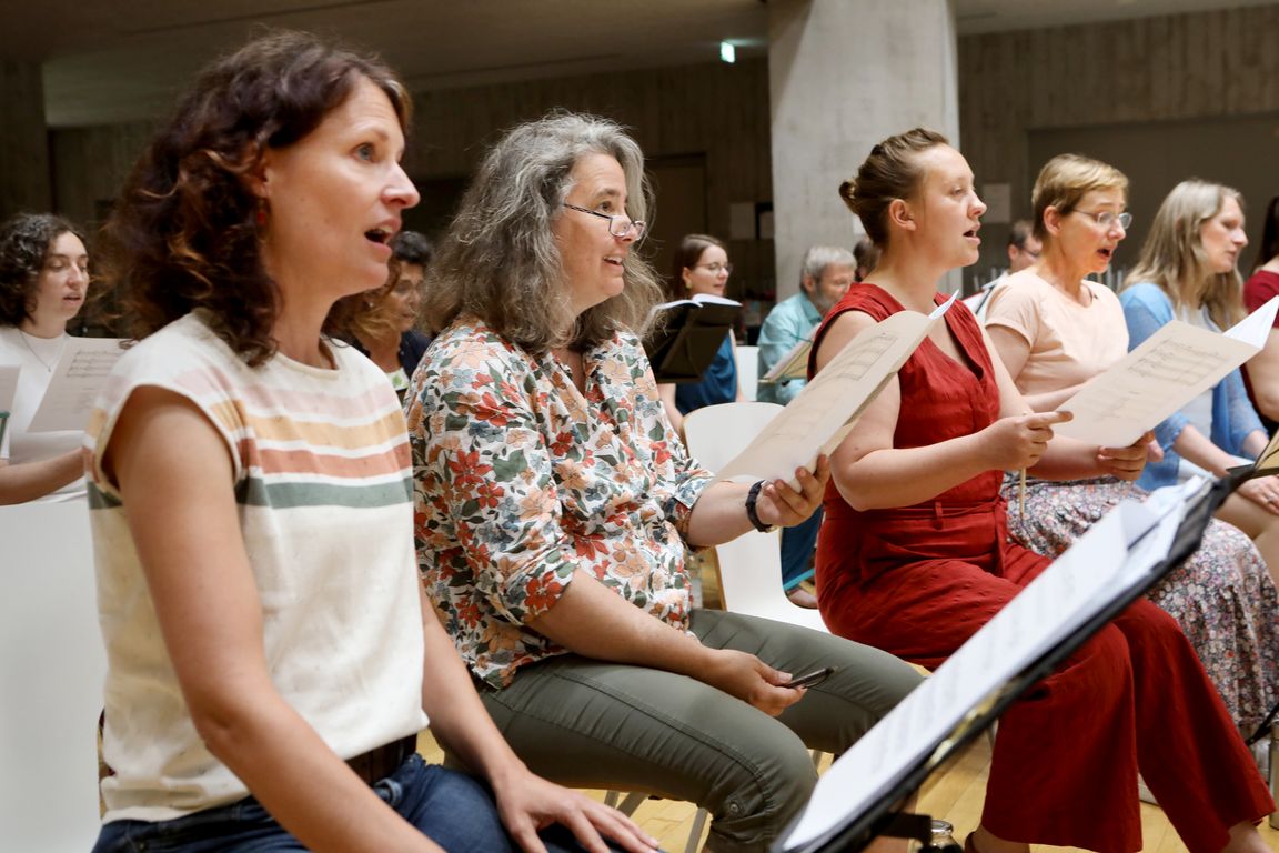 Eine Gruppe von Sängerinnen sitzt im Halbkreis und hält Notenblätter in den Händen. Die Personen sind verschiedenen Alters und wirken engagiert, während sie gemeinsam singen. Im Hintergrund sind weitere Chormitglieder zu erkennen.