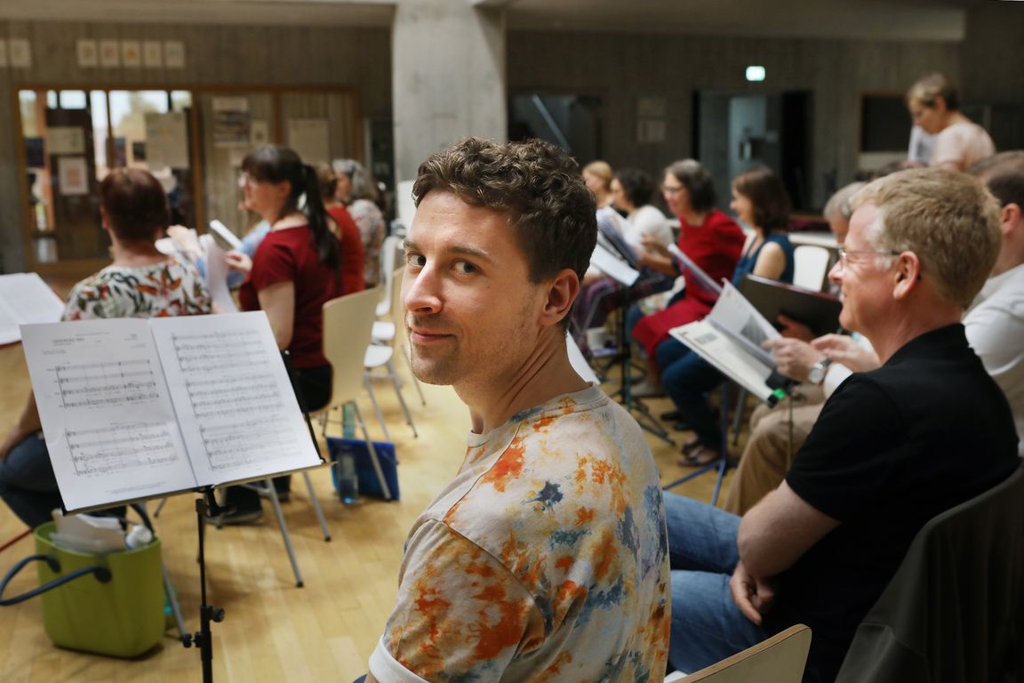 Ein Chorsänger dreht sich während der Probe zur Kamera um und lächelt, während im Hintergrund weitere Mitglieder zu sehen sind. Vor ihm steht ein Notenständer mit aufgeschlagener Partitur. Der Probenraum ist hell, die Stühle sind halbkreisförmig angeordnet.