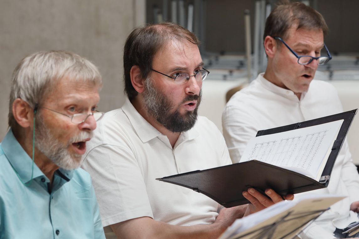 Drei männliche Chorsänger sitzen nebeneinander und singen aus geöffneten Chormappen, ihre Blicke sind dabei nach vorne gerichtet. Die Szene zeigt konzentriertes Singen während einer gemeinsamen Probe.