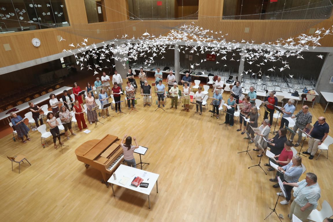 Aufnahme des gesamten Chors in einem großen, lichtdurchfluteten Raum mit Holzfußboden. In der Mitte steht die Dirigentin neben einem Flügel und einem Tisch mit Notenmaterial. Die Sängerinnen und Sänger stehen in weitem Halbkreis um sie herum und halten Noten in der Hand. Über ihren Köpfen hängt eine Installation aus zahlreichen weißen Papierkranichen.