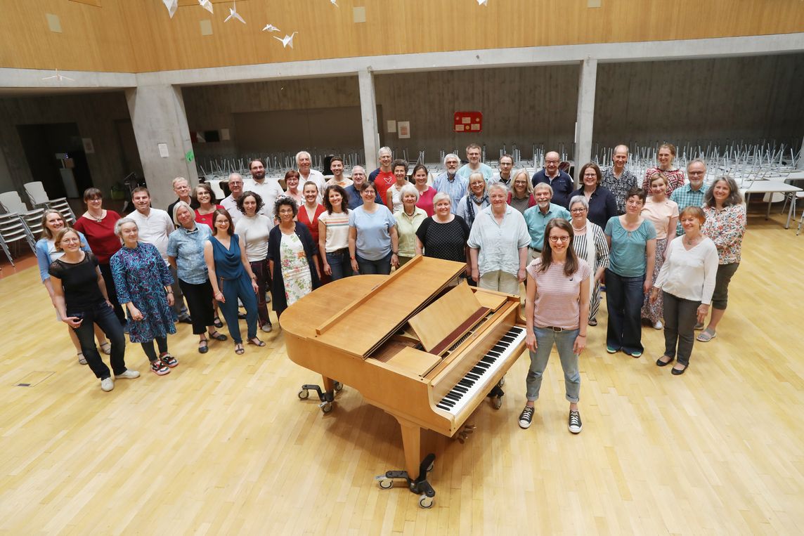 Eine Gruppe von etwa vierzig Sänger:innen steht in der Mitte eines hellen Probenraums halbkreisförmig um einen Flügel herum. Neben dem Instrument steht die Dirigentin. Alle tragen Alltagskleidung und blicken fröhlich in die Kamera. Das Bild zeigt die Vielfalt und Gemeinschaft des Chors.