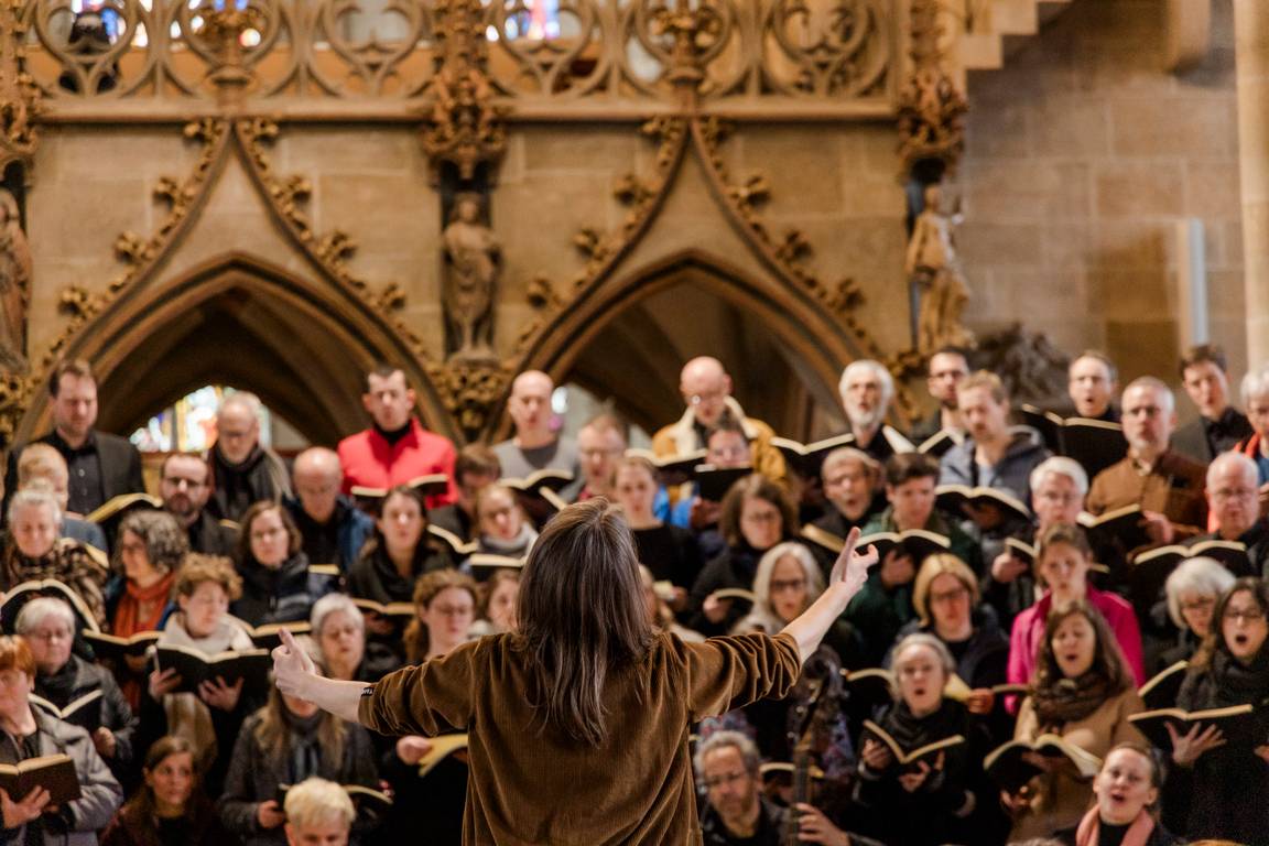 Detail aus der Anspielprobe für die Matthäus-Passion in der Tübinger Stiftskirche: Im Vordergrund steht die Dirigentin mit ausgebreiteten Armen, während im Hintergrund ein Teil des Doppelchores zu sehen ist. Der Fokus der Kamera liegt auf der Dirigentin, die mit dem Rücken zum Betrachter steht. Das Bild zeigt die konzentrierte Zusammenarbeit aller Beteiligten.