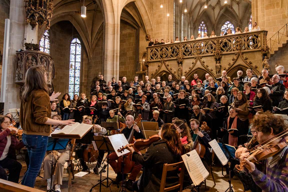 Anspielprobe für die Matthäus-Passion in der Tübinger Stiftskirche. Im Vordergrund sind die Dirigentin und das Orchester zu sehen, im Hintergrund der Chor, der in mehreren Reihen vor dem Altar steht. Die Sänger:innen halten schwarz eingebundene Partituren und konzentrieren sich auf ihren Einsatz. Oberhalb des Altars auf dem sogenannten Lettner stehen ungefähr vierzehn Kinder im Grundschulalter, die den Cantus firmus im Eingangschor mitsingen. Die gotische Architektur der Kirche bildet einen eindrucksvollen Rahmen für den musikalischen Auftritt.